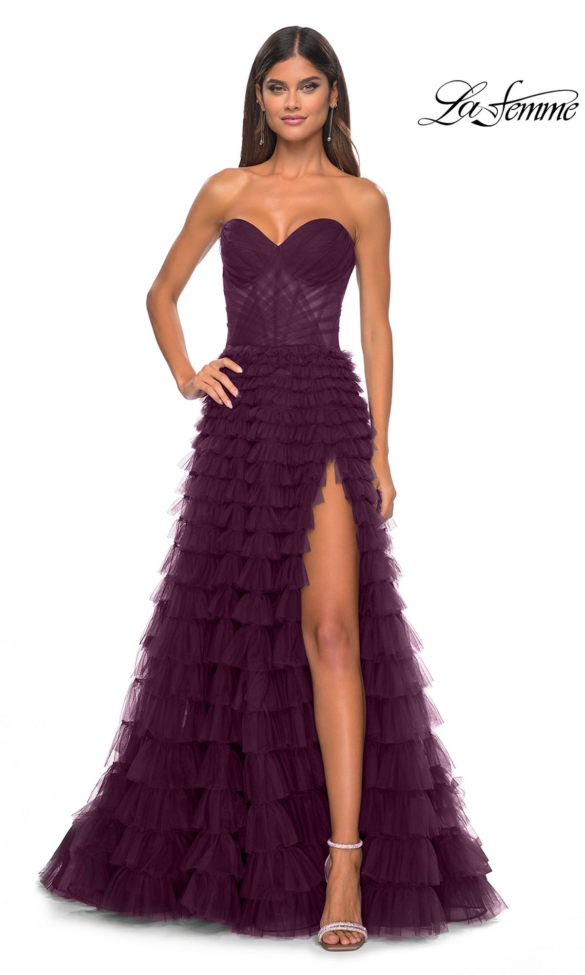 A woman poses against a white background in the La Femme Strapless Long Ruffled Prom Dress 32283—a deep purple A-line gown with a high slit and layered tulle—paired with high-heeled sandals for an elegant look.
